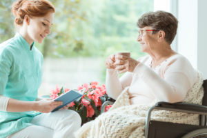 A caregiver reads to an elderly woman sitting in a chair and holding a mug