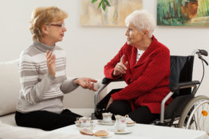 A middle-aged woman talks to an elderly woman in a wheelchair over drinks at a coffee table